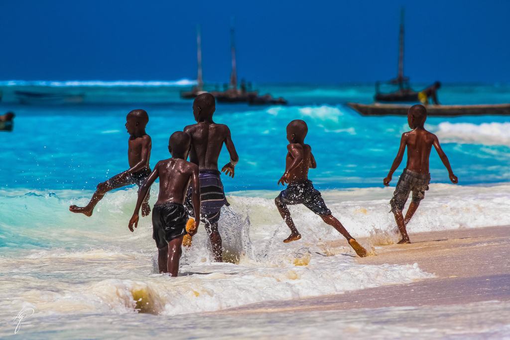 Fünf Jungen laufen durch die Brandung, dahinter Segelboote auf türkisem Wasser