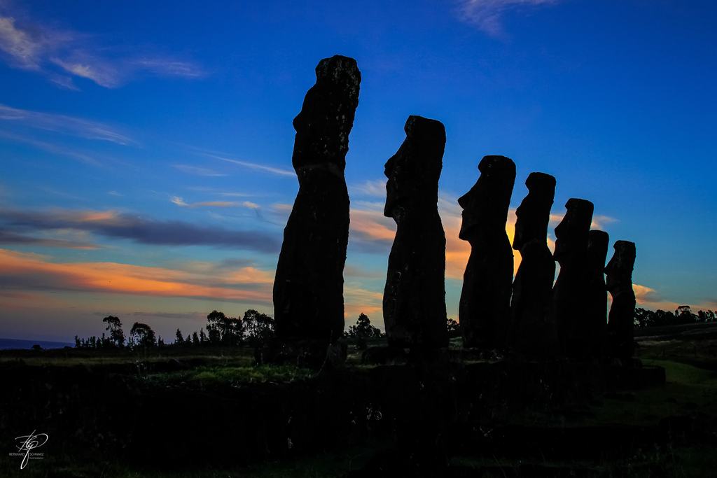 Sieben Steinfiguren stehen als Reihe von Silhouetten im Abendlicht