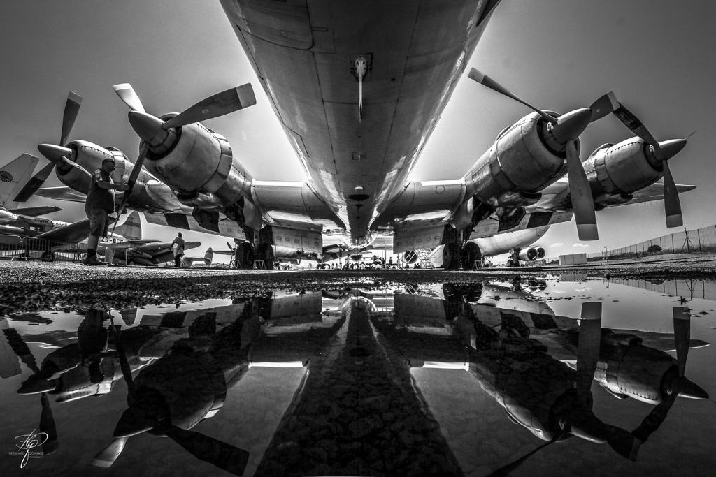Schwarzweiß: ein viermotoriges Propellerflugzeug spiegelt sich in einer Pfütze